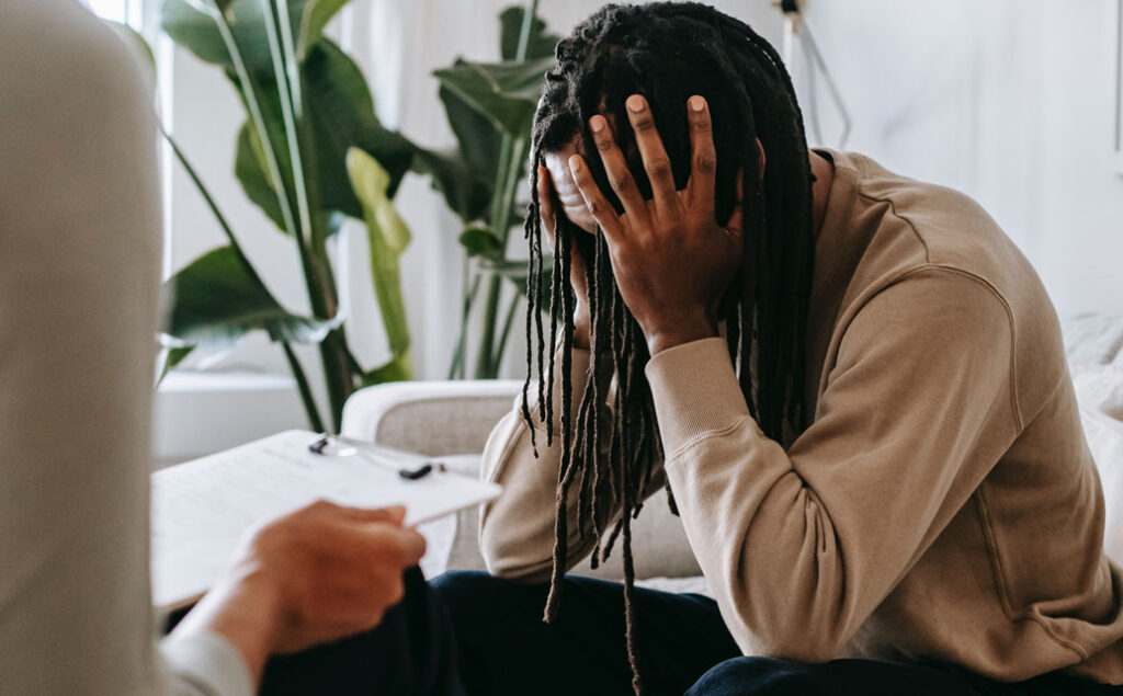 An African American man holding his head, face covered, in presumably a doctors office