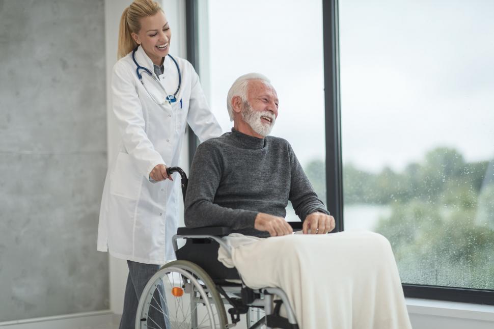 Alt: A smiling AFAB doctor pushing an elderly AMAB patient in a wheelchair, both appearing happy.