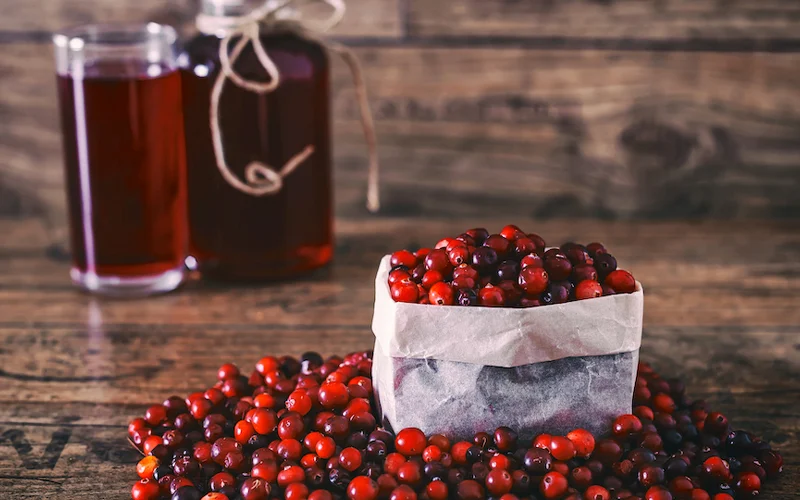 An image of a box of cranberries, with berries on the table near the box, and two glasses of cranberry juice in the background.