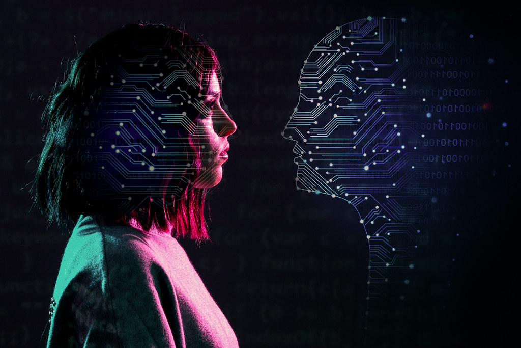 A woman with neck length hair and an overlay of transparent circuits looks at a facade of a human, implied to be AI, which is filled in with those same transparent circuits, dark background, blue/red lighting.