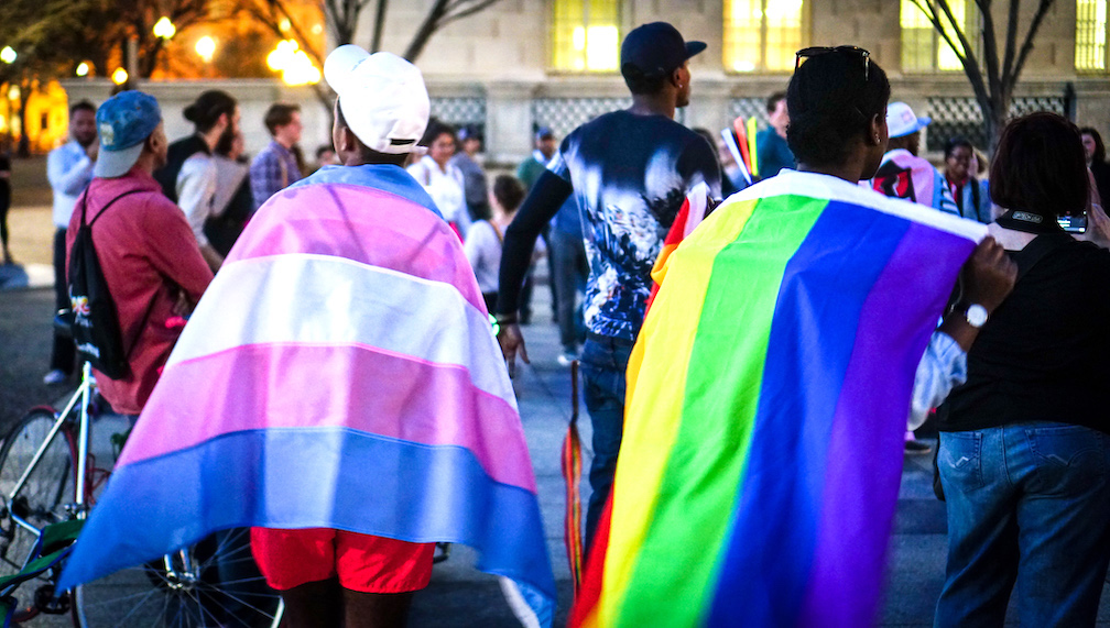 A group of individuals gathers outdoors at dusk, engaging in a social or advocacy event. Two participants in the foreground are wearing pride flags—one displaying the transgender flag and the other the rainbow LGBTQ flag—draped over their shoulders. The scene is lively, with bicycles and other attendees visible in the background, set against the backdrop of an urban environment.