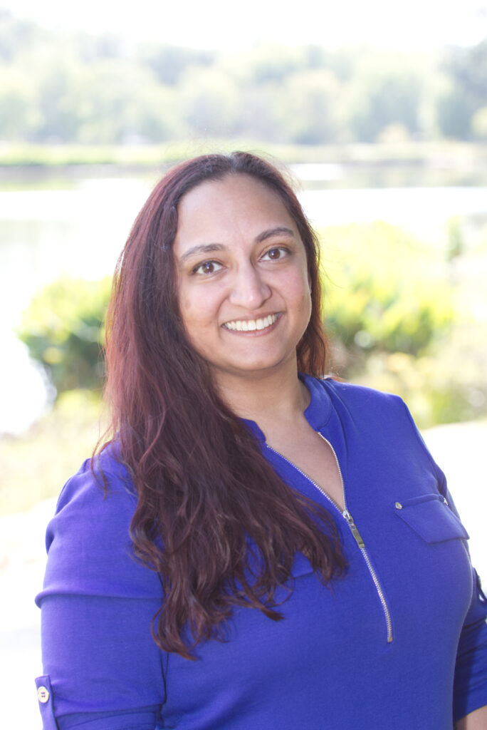 A Professional headshot of Grace R, in a blue top with a background of a park. Grace has roughly shoulder length dark reddish-brown hair, and is facing the camera, smiling.