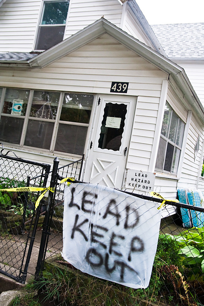 A house with white siding cordoned off by caution tape. A hand-painted sign on the fence reads "LEAD KEEP OUT," and another sign near the door says "LEAD HAZARD KEEP OUT," indicating environmental contamination and health hazards.